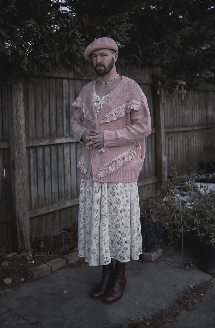Man wearing a pink cardigan, lace top, and long floral skirt standing by a wooden fence.