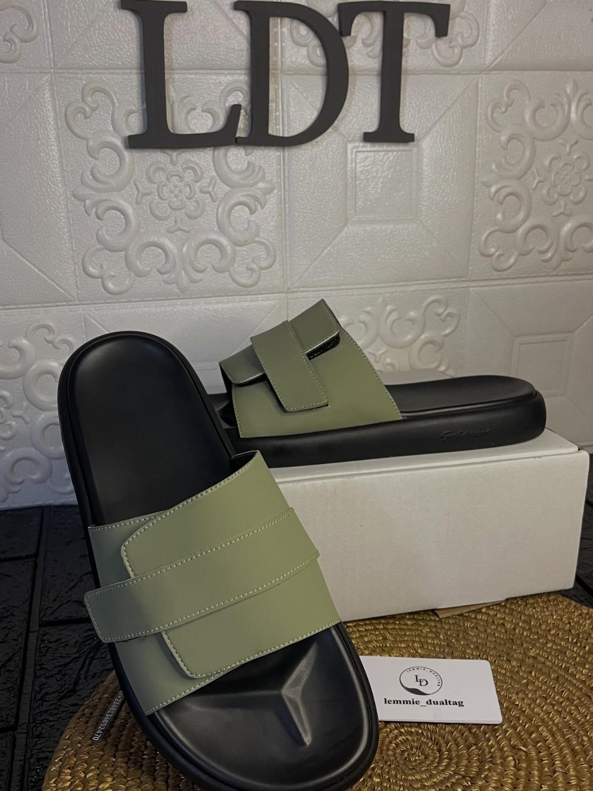 Sage green slide sandals with wide straps resting on a white box and woven mat.