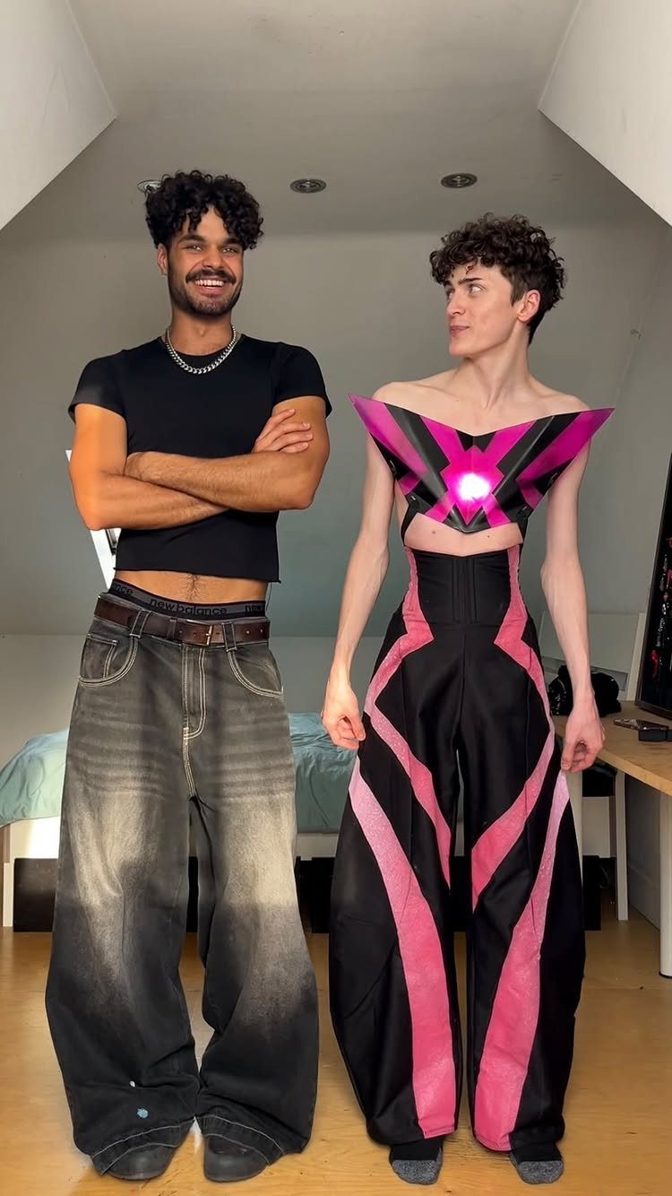 Two people posing in stylized, gender-neutral clothing featuring oversized denim and geometric pink patterns.