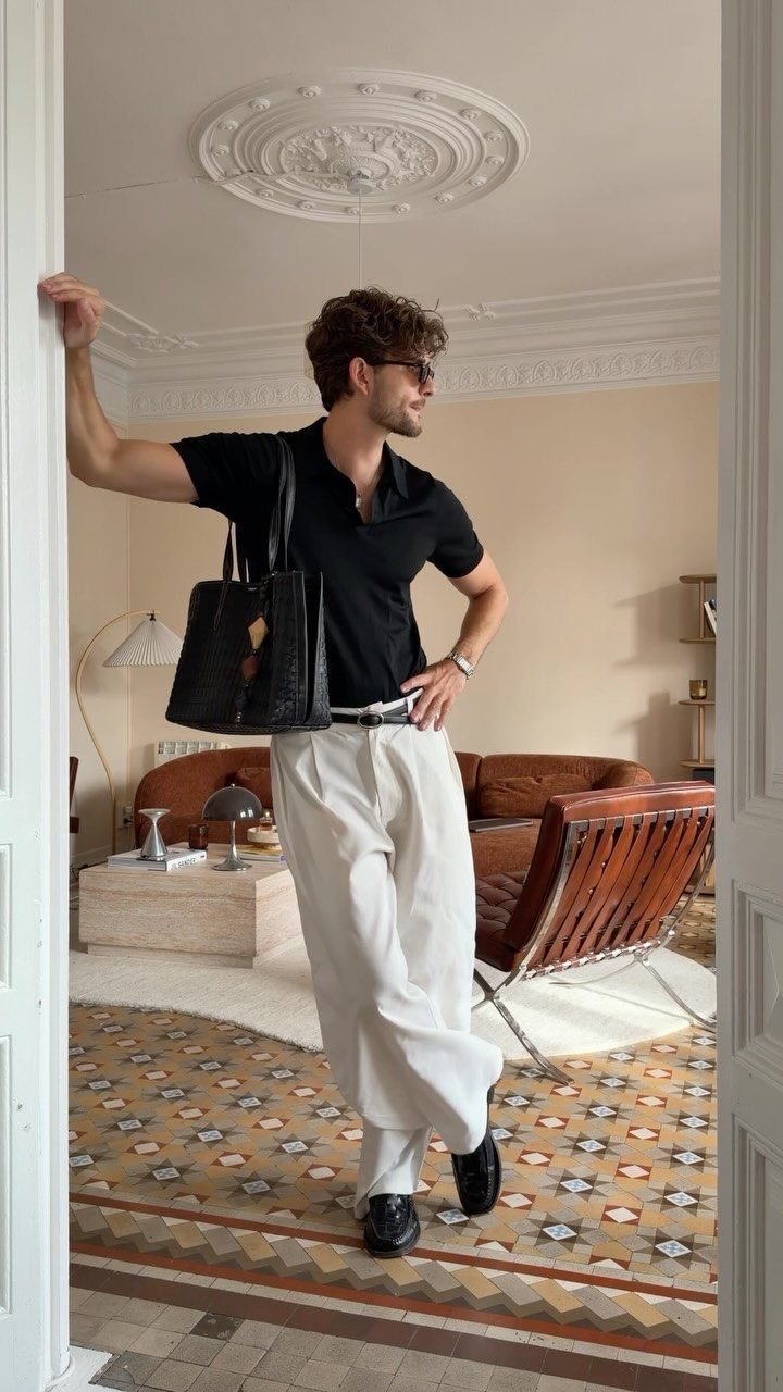 A man in a black polo and white wide-leg trousers posing in a stylish living room.