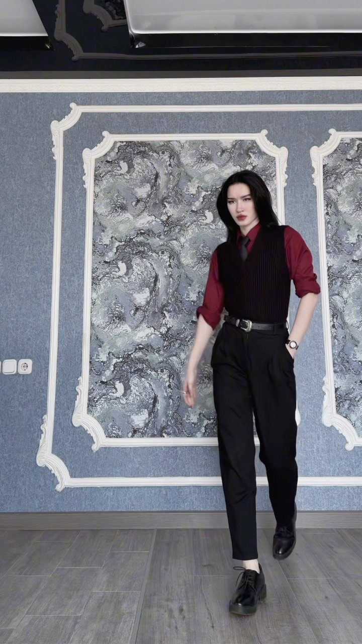 A person wearing a black sweater vest, maroon shirt, and black trousers posing against a blue wall.