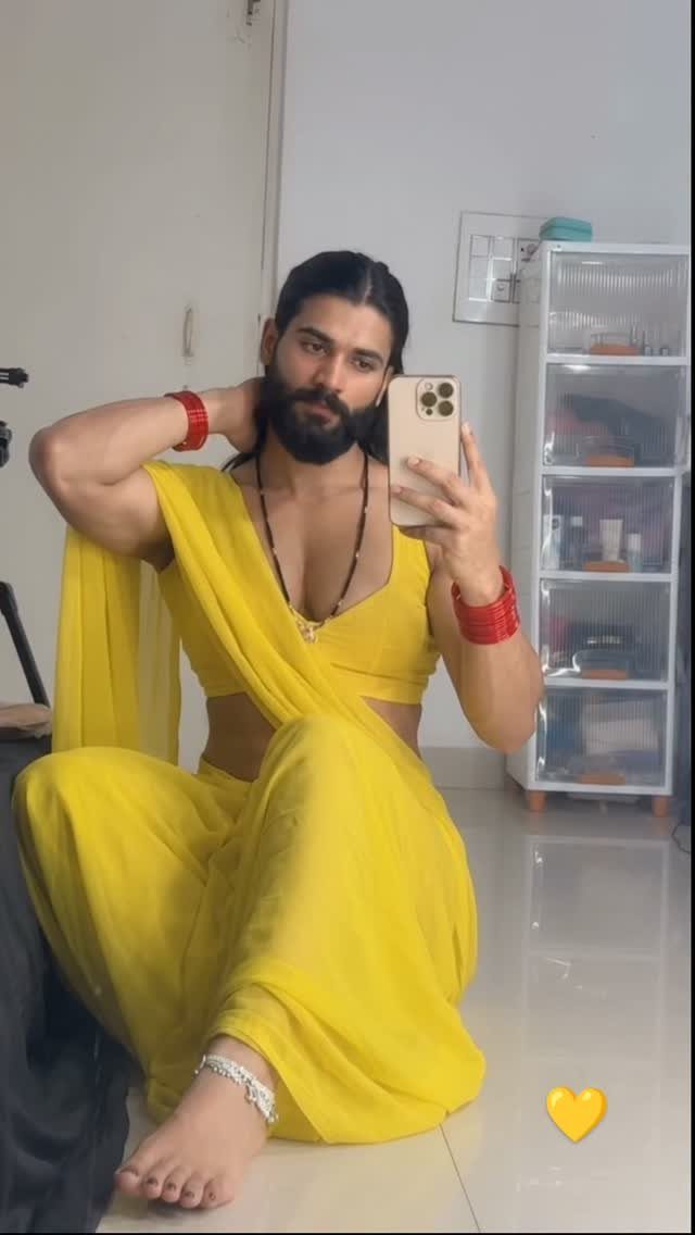 A bearded man posing in a bright yellow saree and red bangles while taking a selfie.
