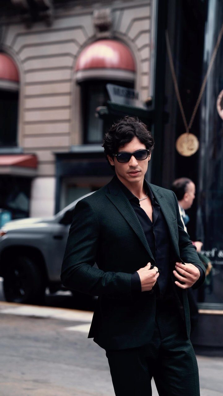 Man in a dark tailored suit and sunglasses standing on a city street.