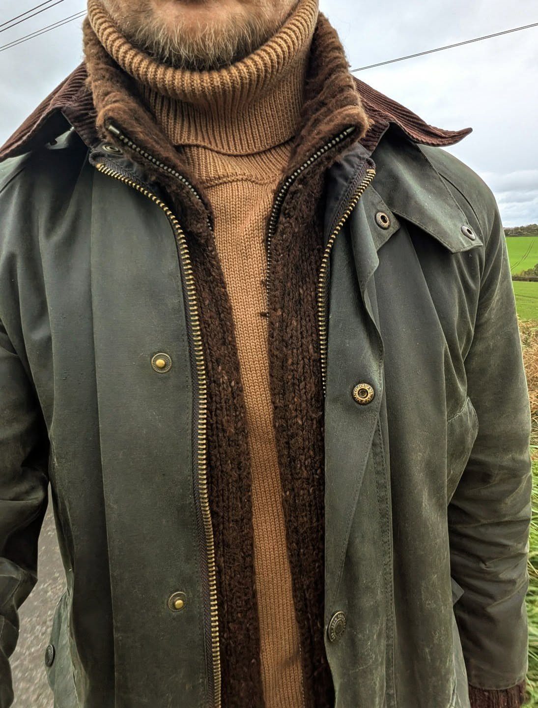 A man wearing a tan turtleneck, brown cardigan, and olive green waterproof coat outdoors.