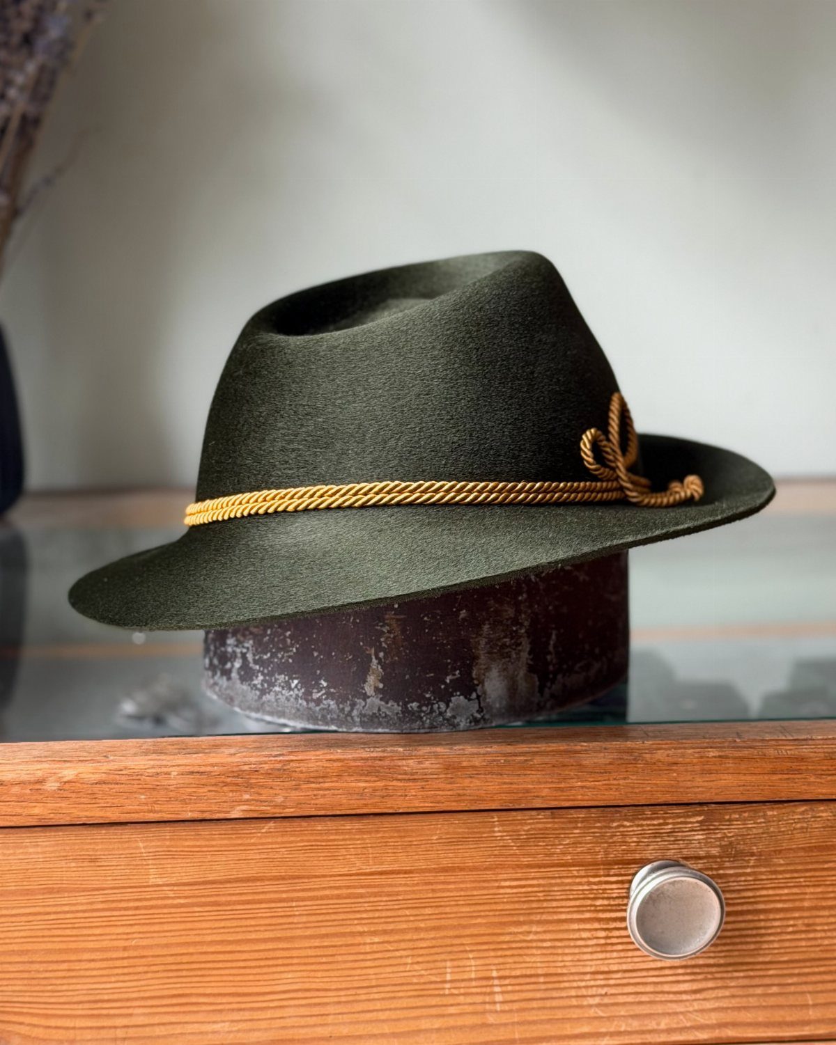 A dark green felt Tyrolean hat with a gold braided cord band resting on a stand.