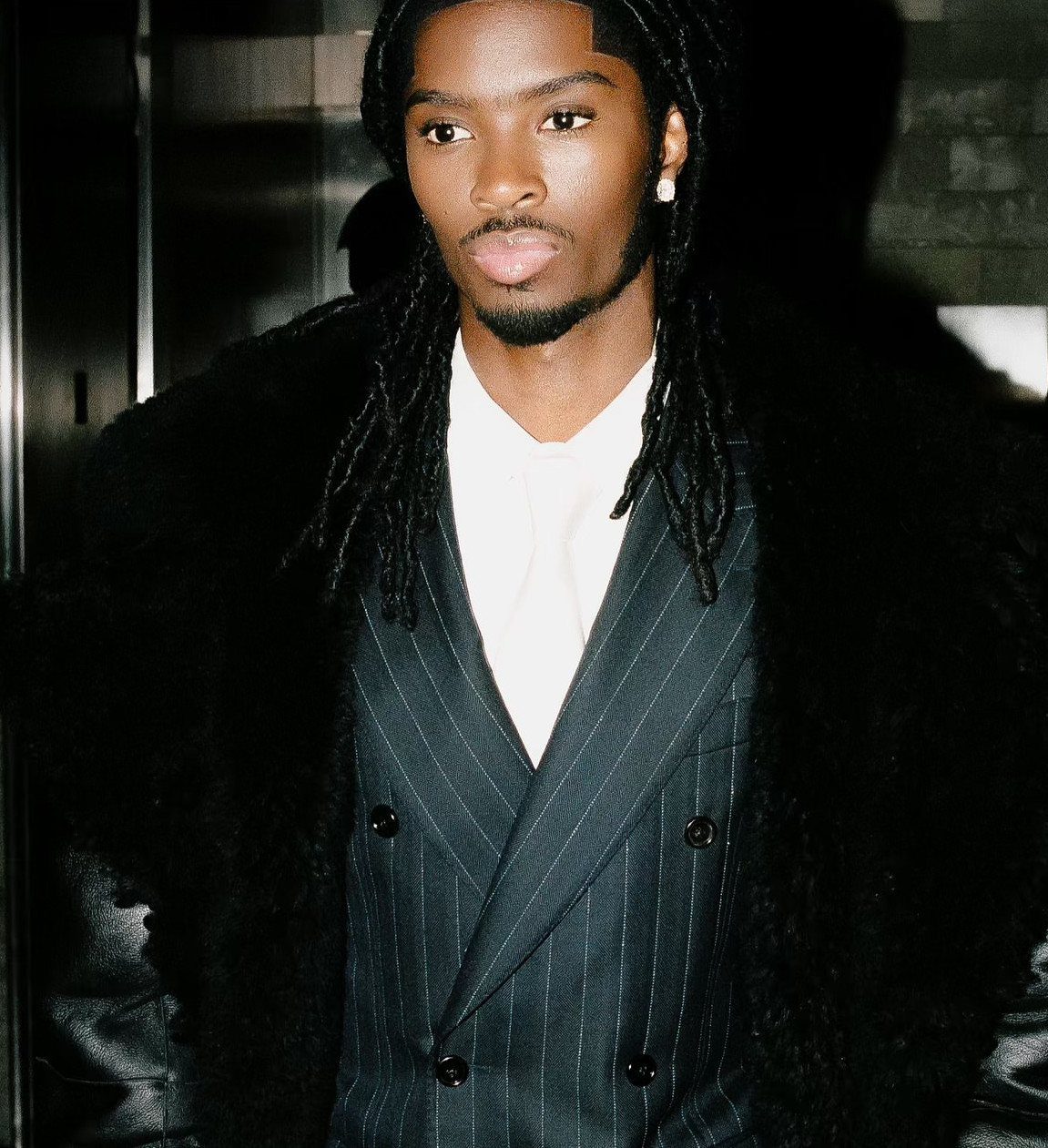 A male model with dreadlocks wearing a pinstripe waistcoat and a black textured coat.