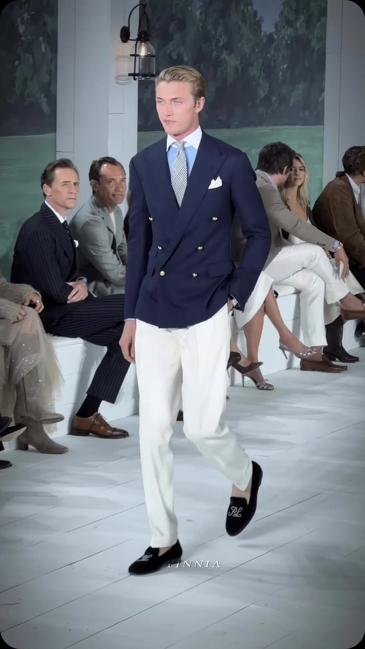 Blonde male model walking a runway in a navy blazer and white trousers.