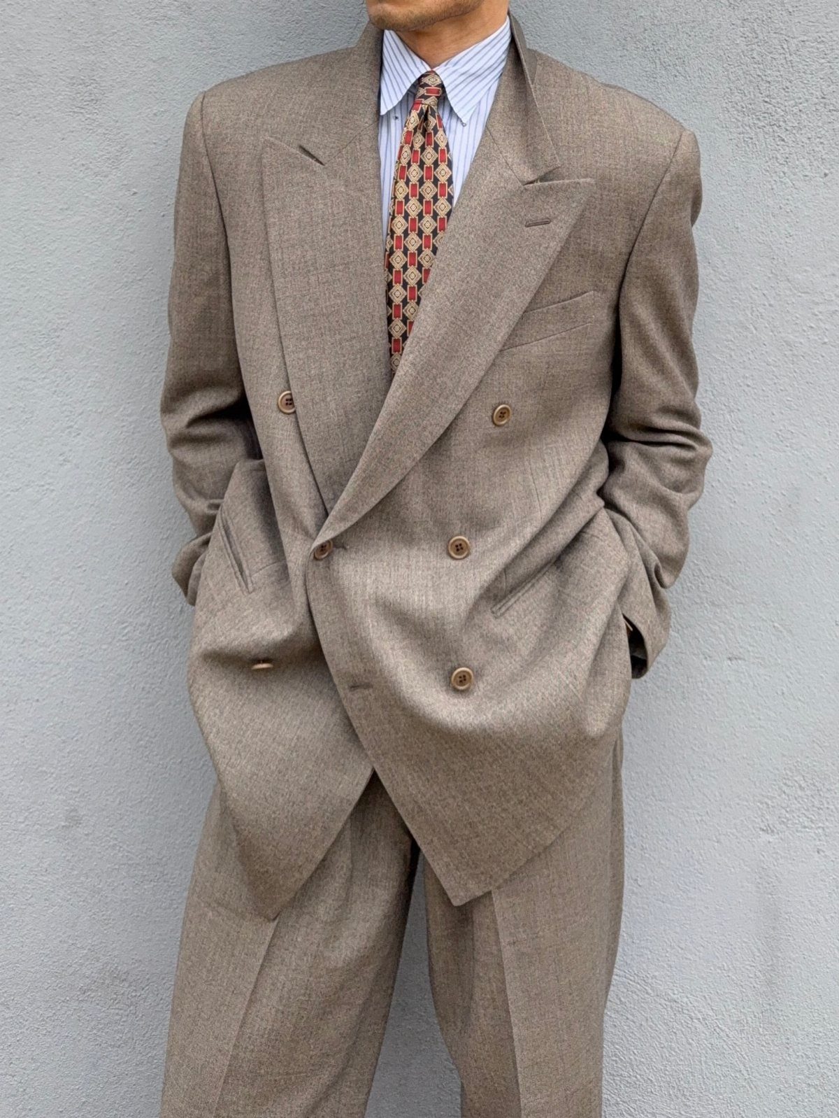 A man wearing a vintage oversized tan double-breasted suit with a patterned tie against a grey wall.