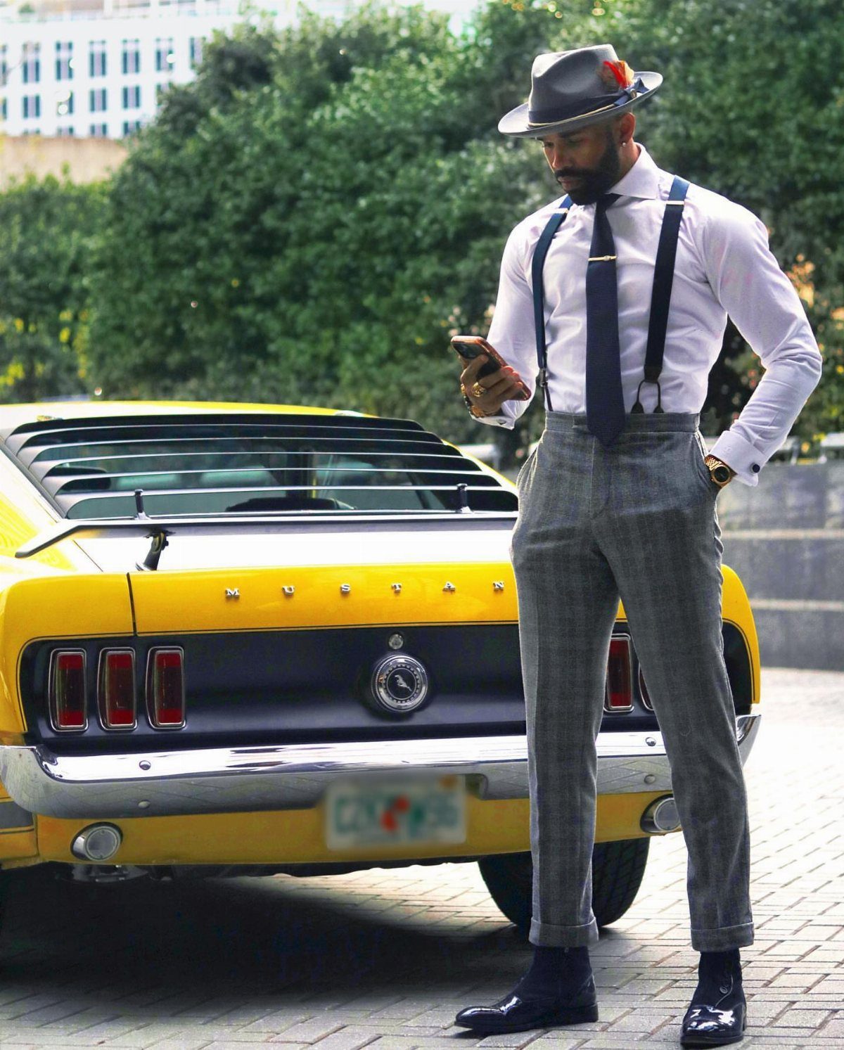 Man in white shirt, dark tie, and grey patterned trousers with suspenders standing by a yellow car.