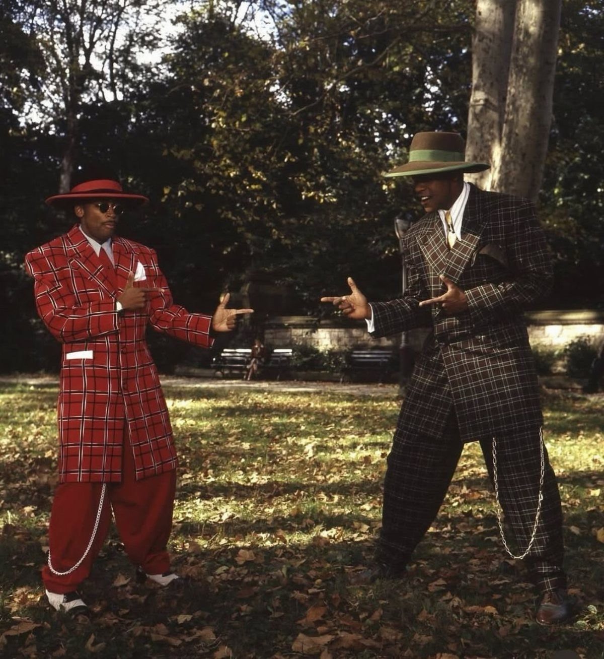 Two men posing in oversized, patterned zoot suits in a park with fallen leaves.