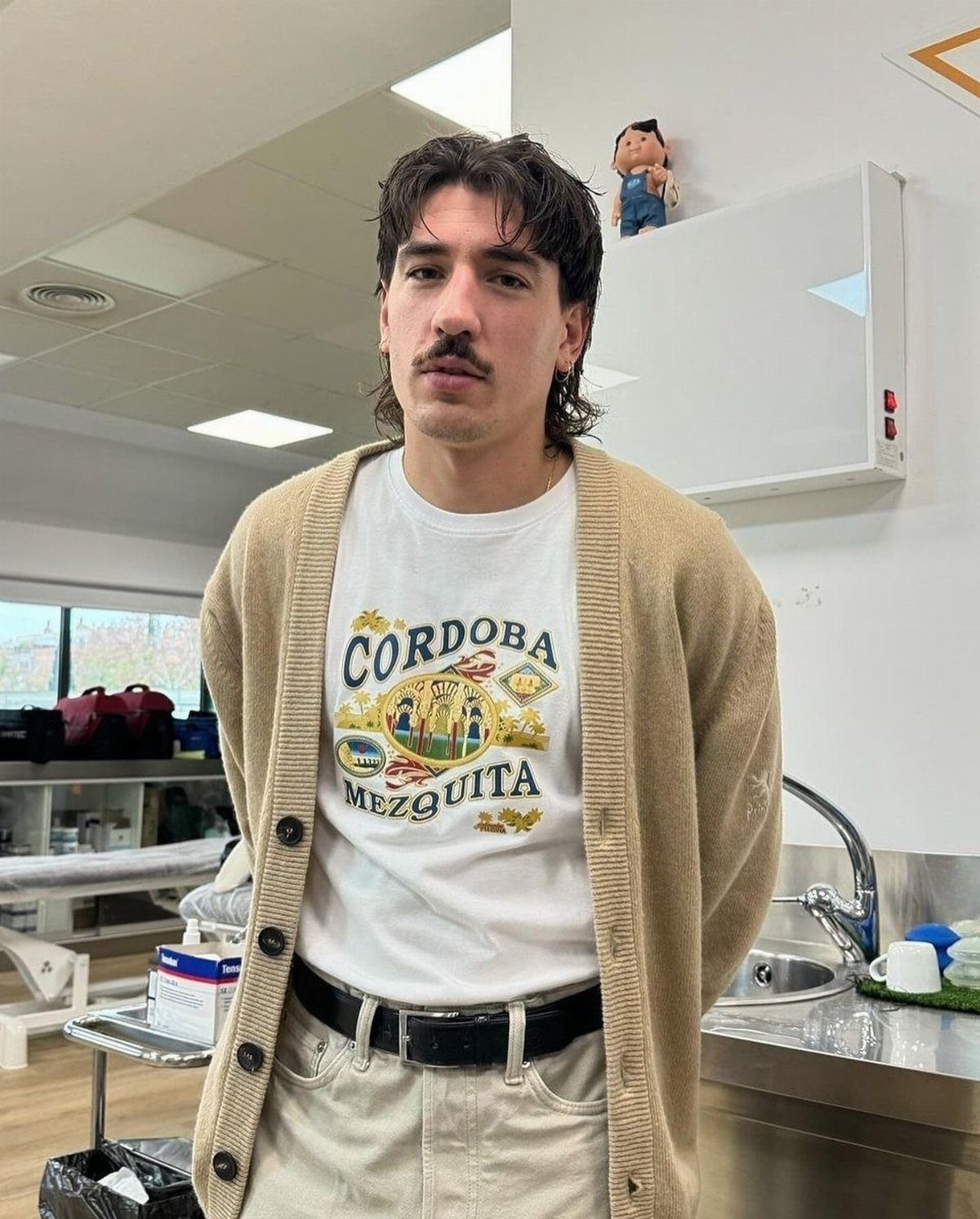 Man with a mustache wearing a white graphic t-shirt and a beige cardigan indoors.
