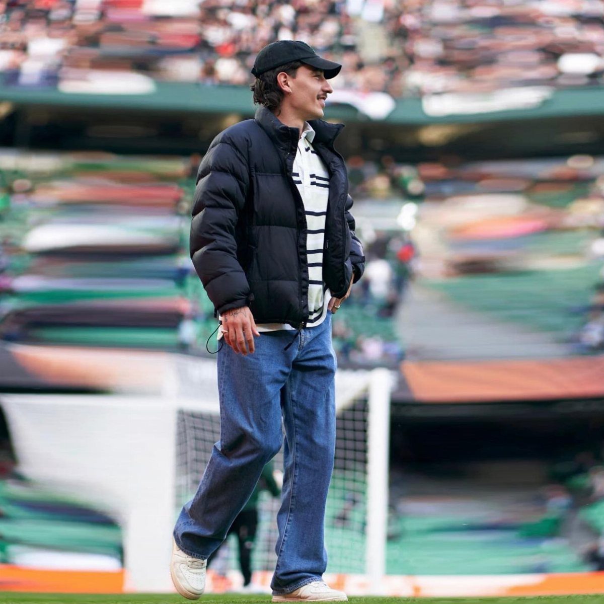 A man in a black puffer jacket and blue jeans walking on a football field.