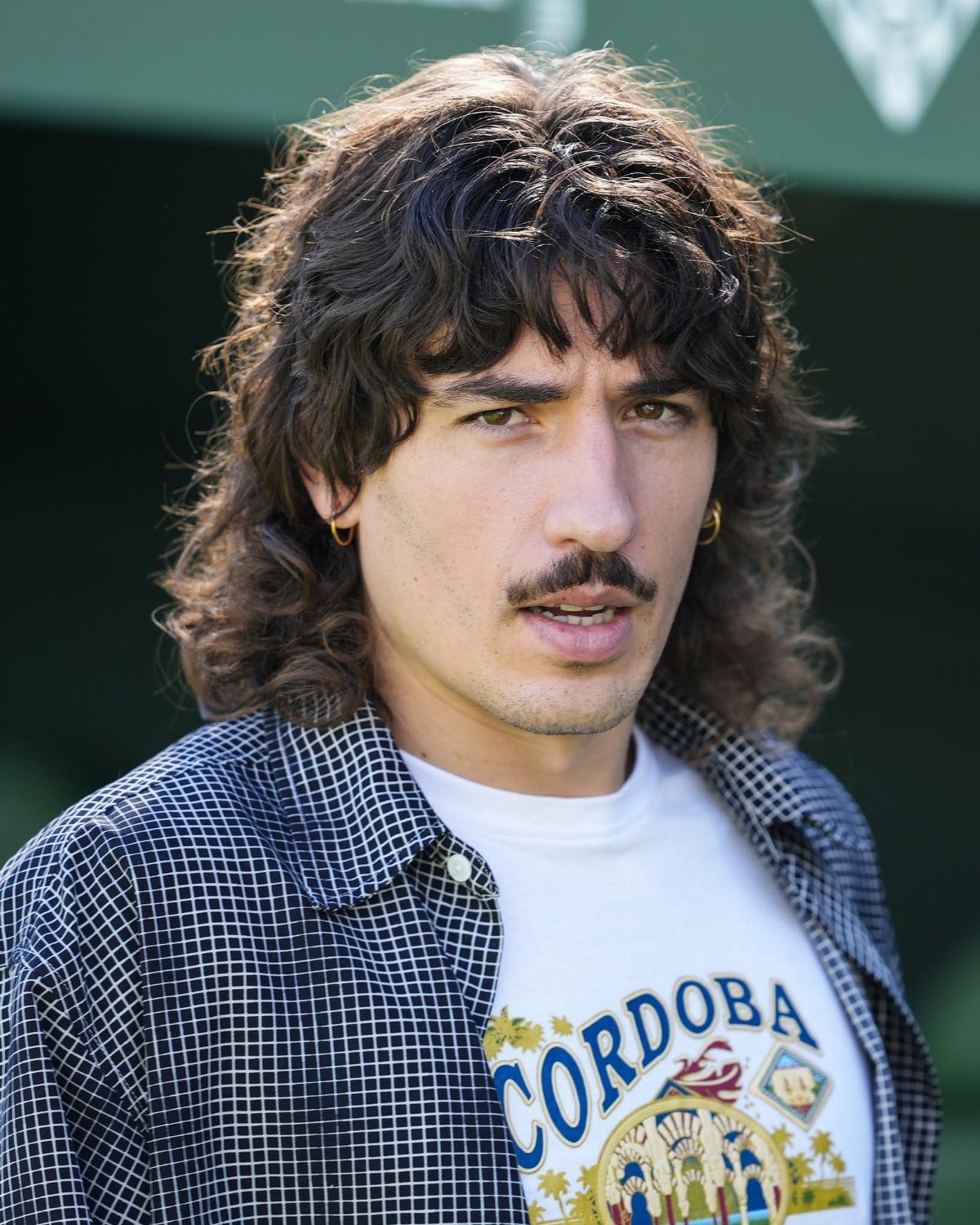 A man with wavy hair and a mustache wearing a checkered shirt and graphic t-shirt.