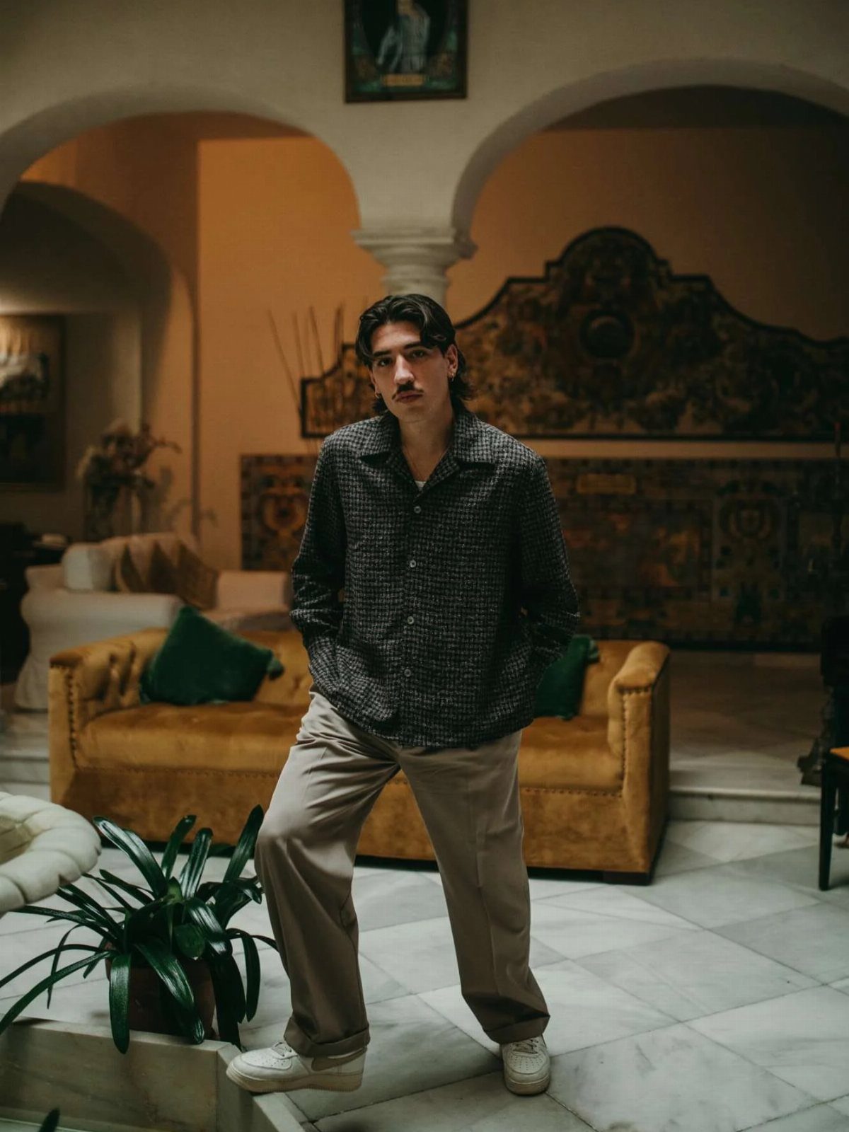 A young man in a patterned shirt and tan trousers posing in a room with arches and a yellow sofa.