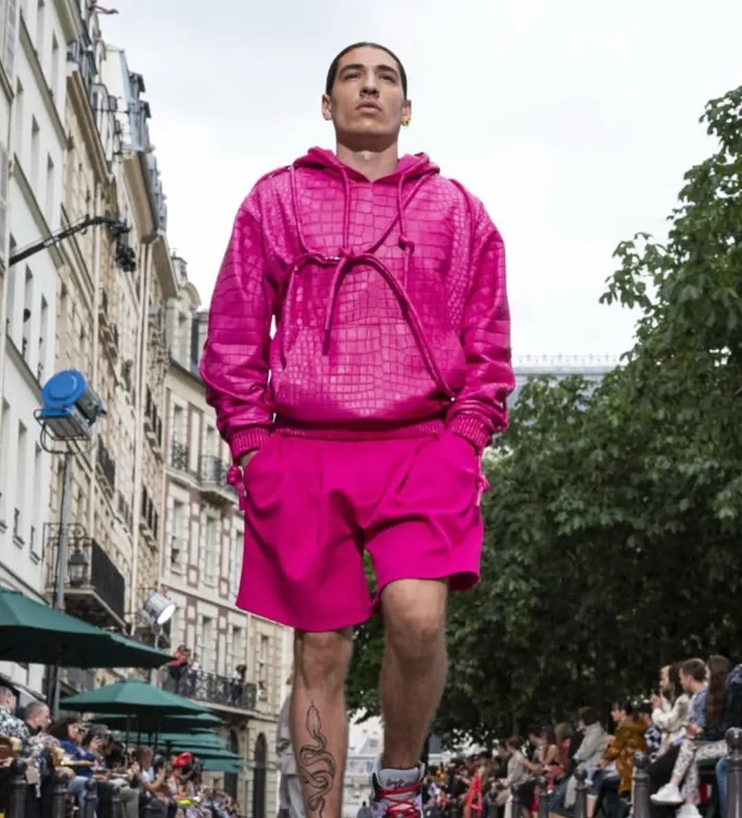 A man walking in a bright fuchsia croc-embossed hoodie and matching shorts through a city street.
