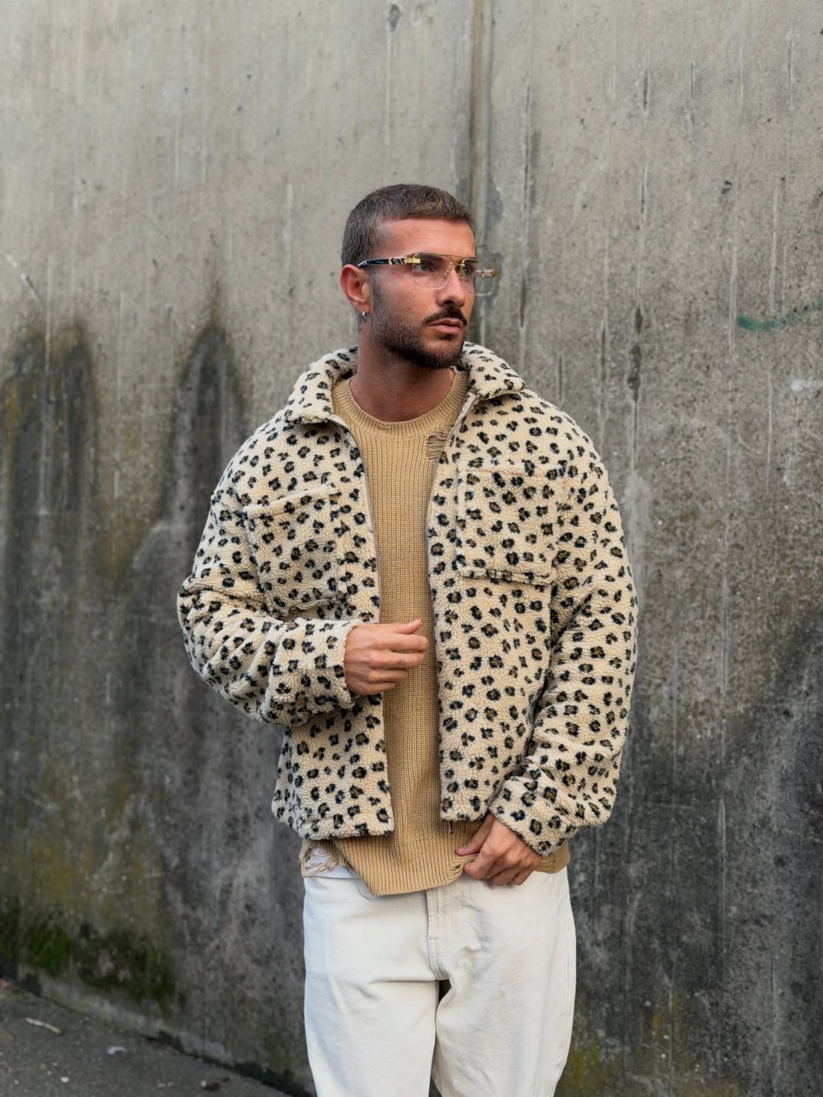 Man in leopard print fleece jacket and beige sweater posing against a concrete wall.