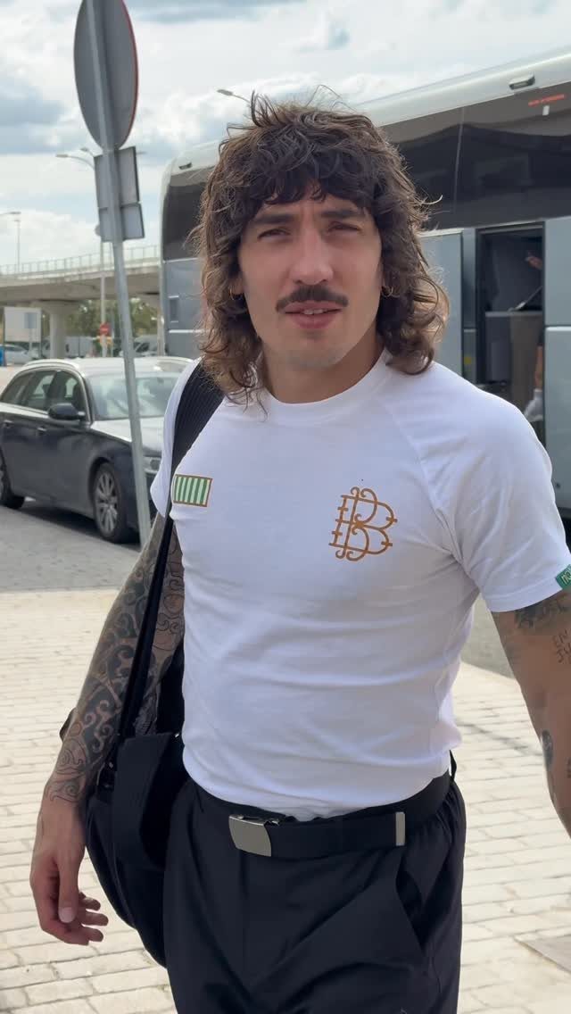 Man with long hair and mustache wearing a white logo t-shirt and black trousers outdoors.