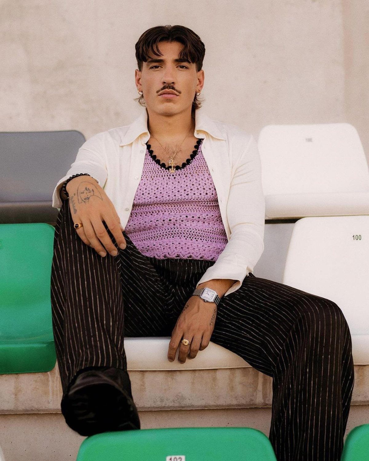 Man in purple crochet top and pinstriped trousers sitting on stadium seats.