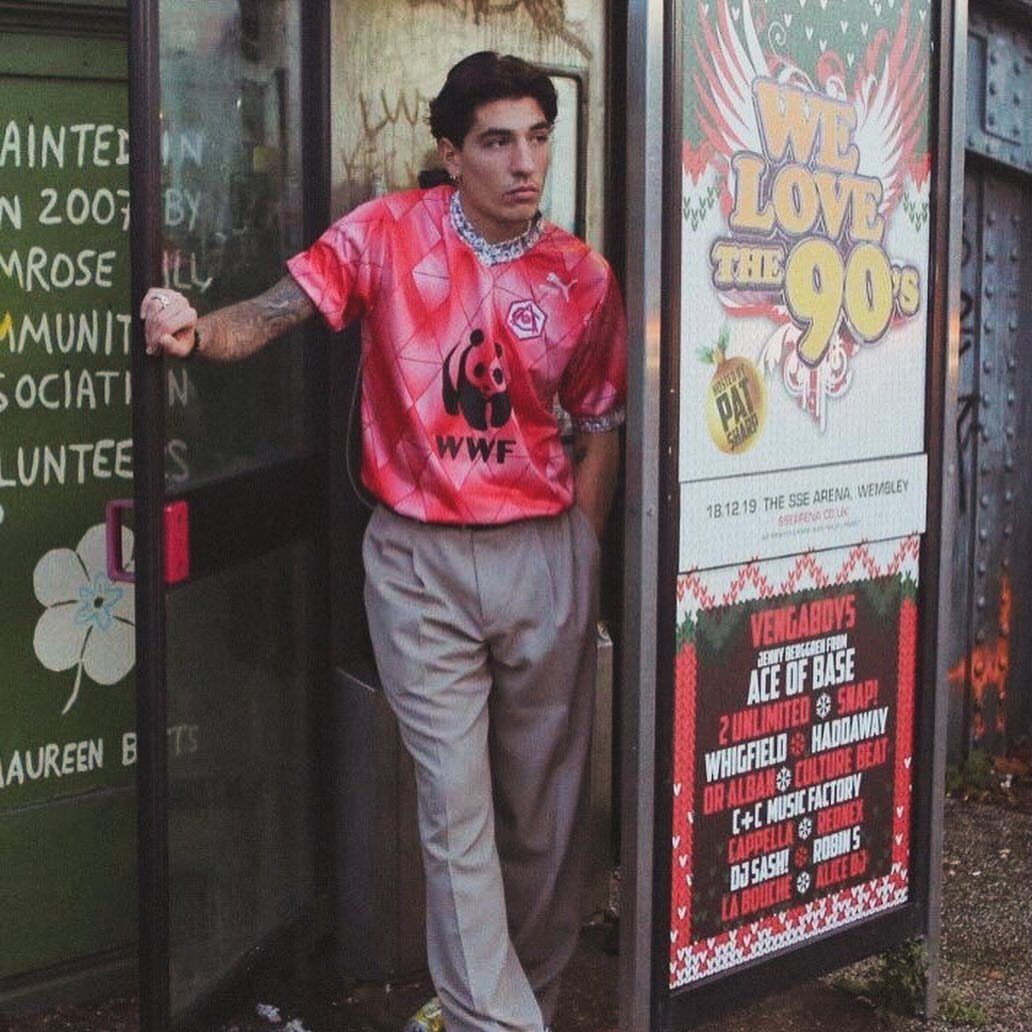 A man in a pink WWF soccer jersey and grey trousers leaning against a doorway.