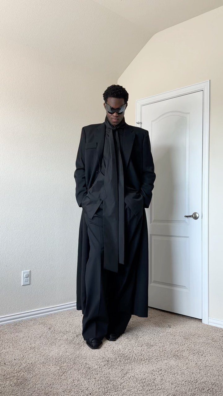 A person posing in an all-black high fashion outfit with a long coat and sunglasses.