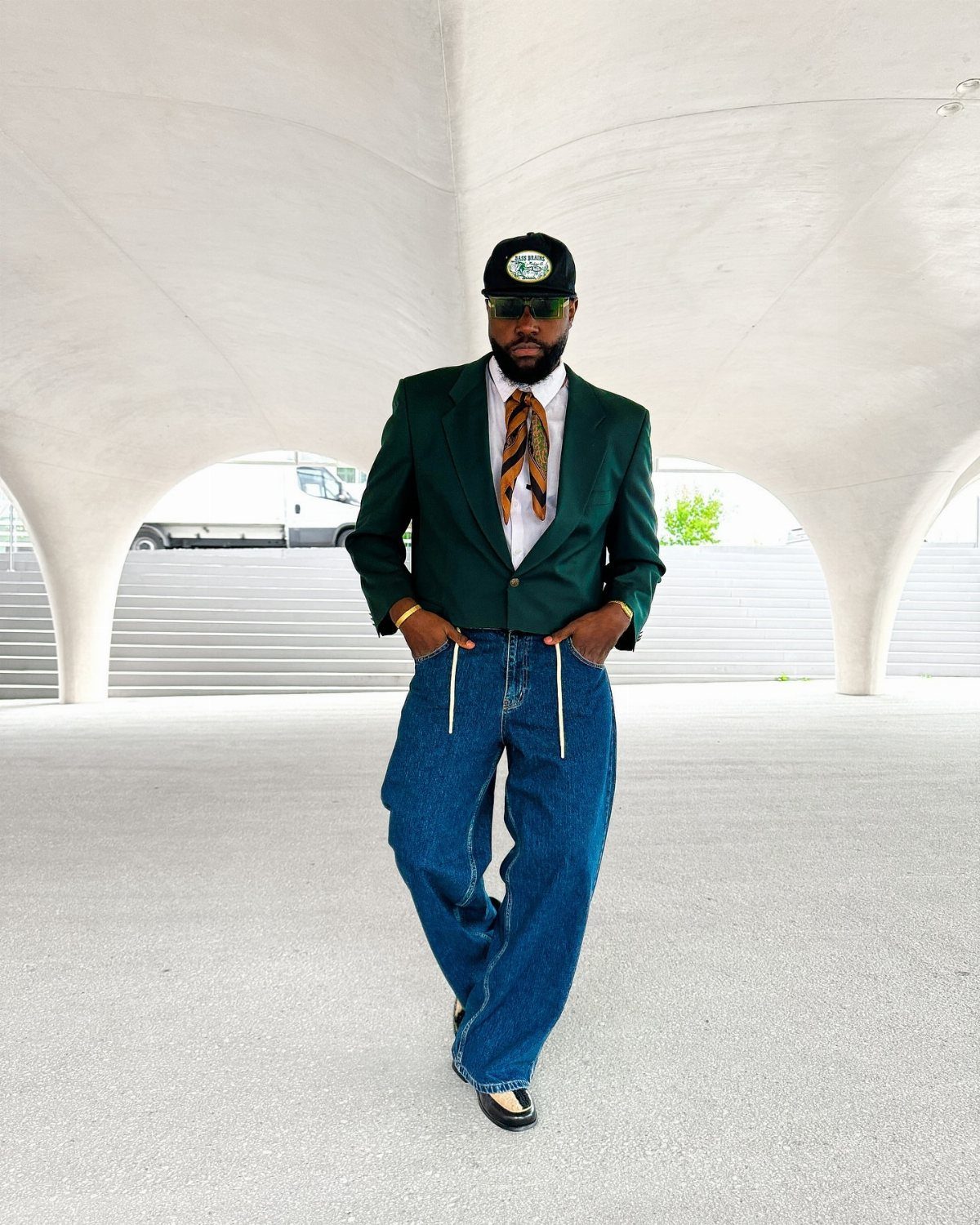 A man in a green blazer, patterned silk tie, and baggy jeans posing outdoors.
