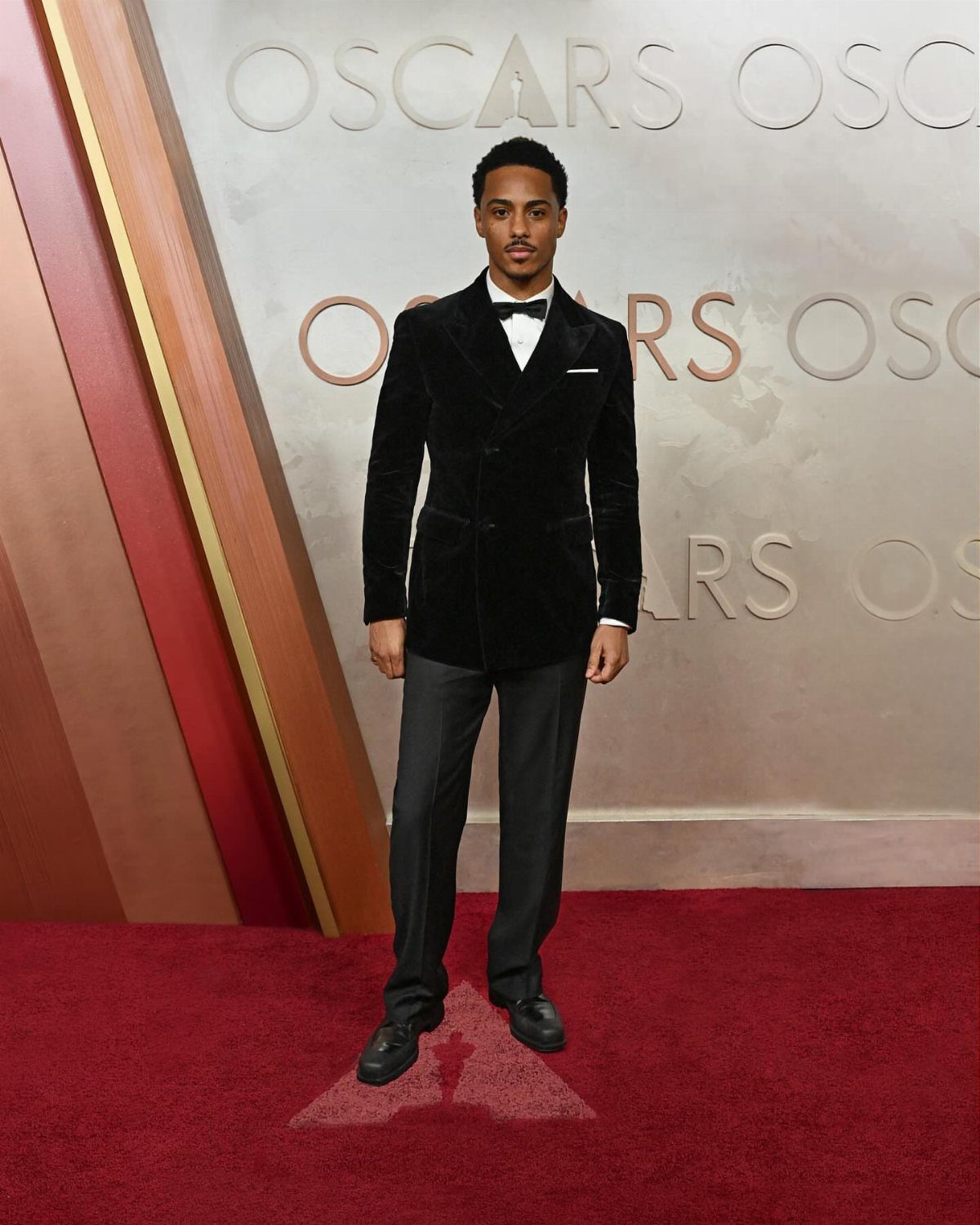 A man in a black velvet tuxedo posing on a red carpet at the Oscars.