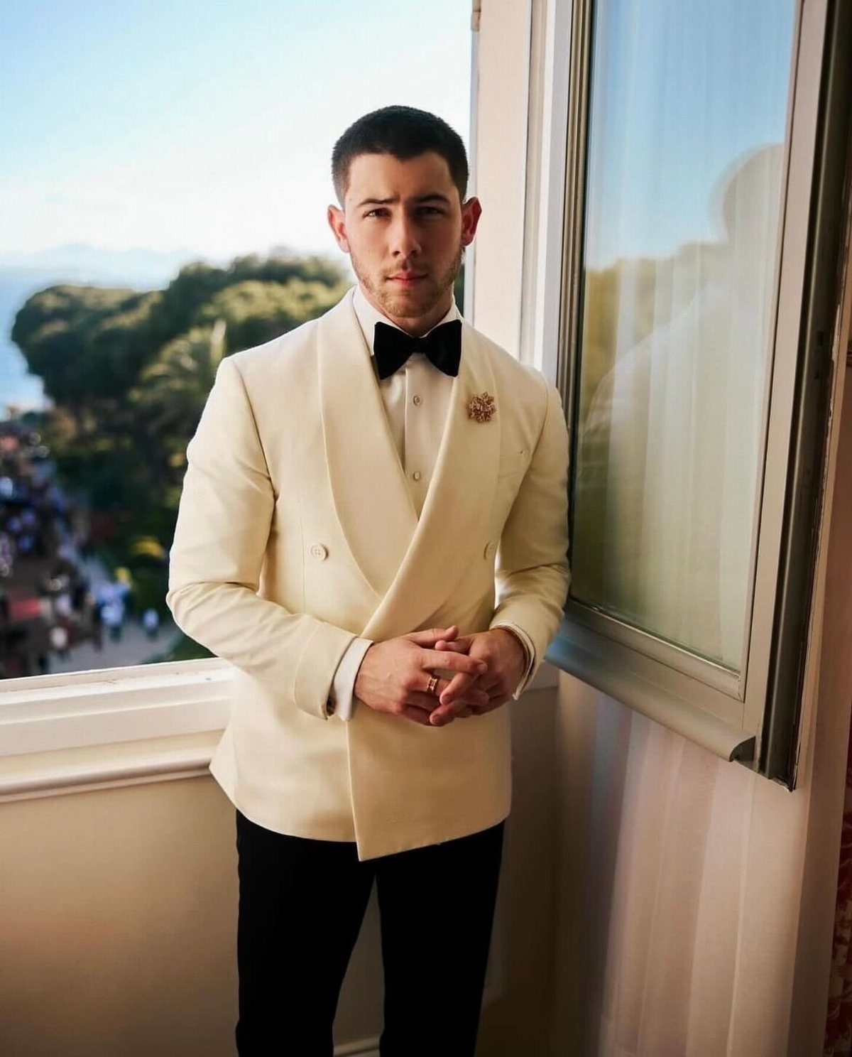 Man in a cream tuxedo jacket and black bowtie standing on a balcony overlooking a coast.