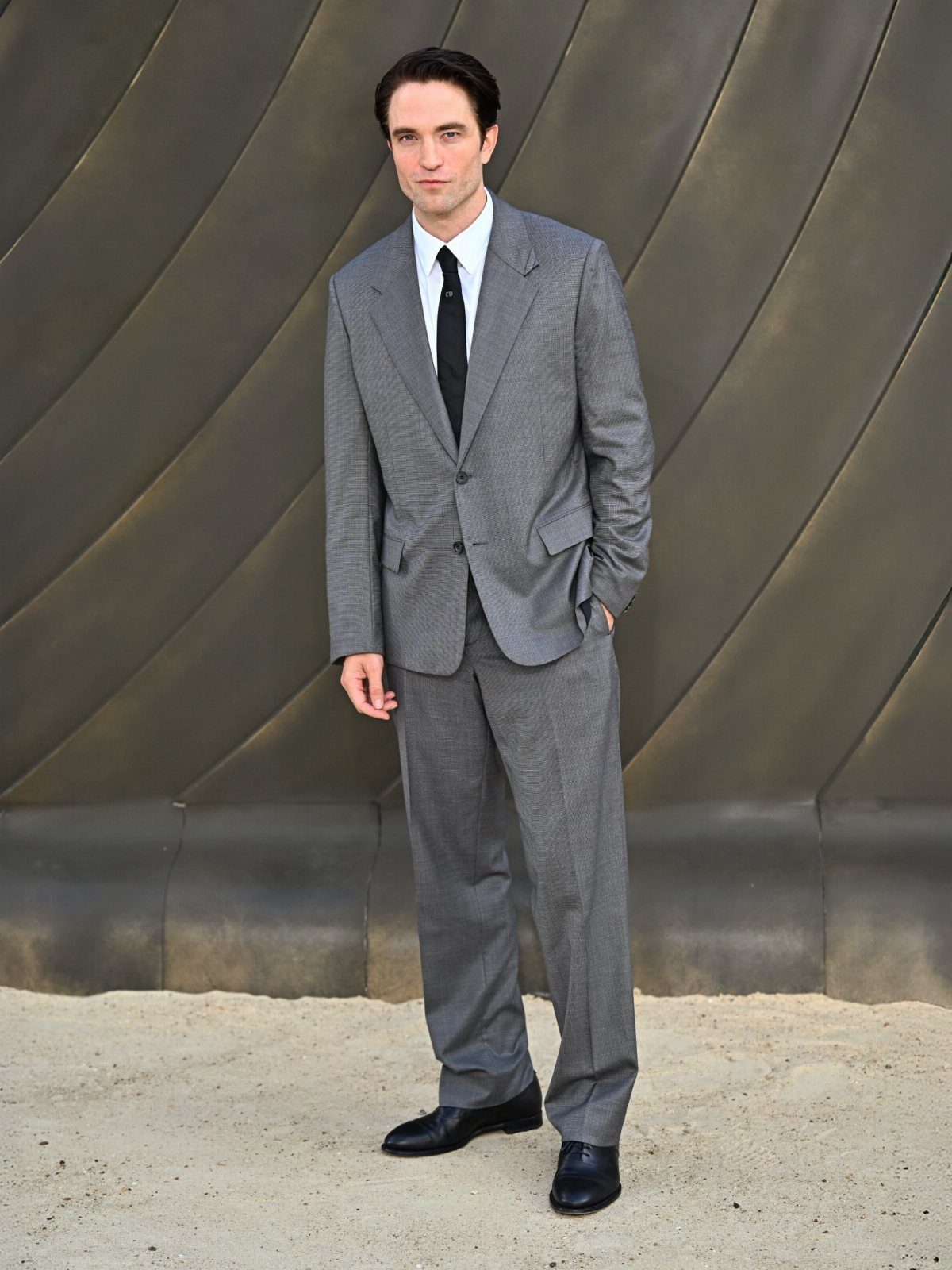 A man in a grey patterned suit and black tie posing against a metallic background.