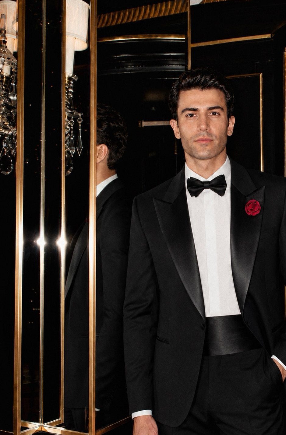 Man posing in a black tuxedo with satin lapels, a bowtie, and a red rose boutonniere.