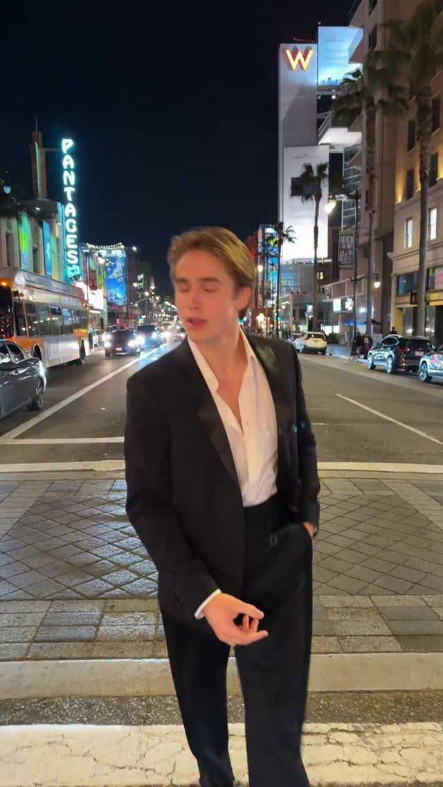 Young man in a black suit and white shirt standing on a city street at night.