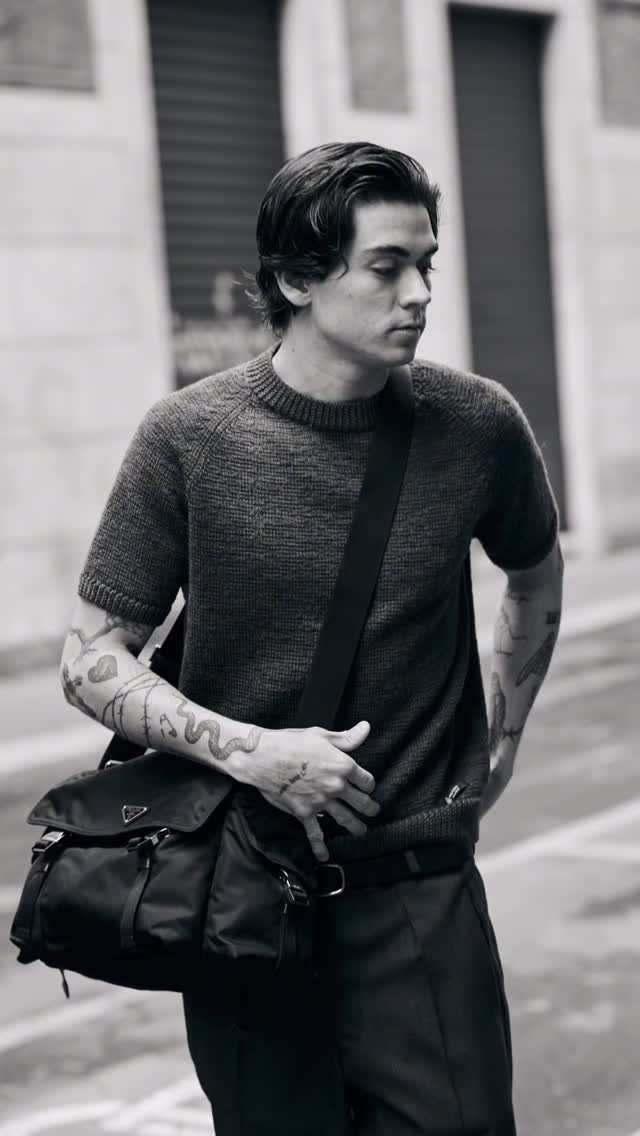 A tattooed man in a knit sweater carrying a black Prada crossbody bag outdoors.