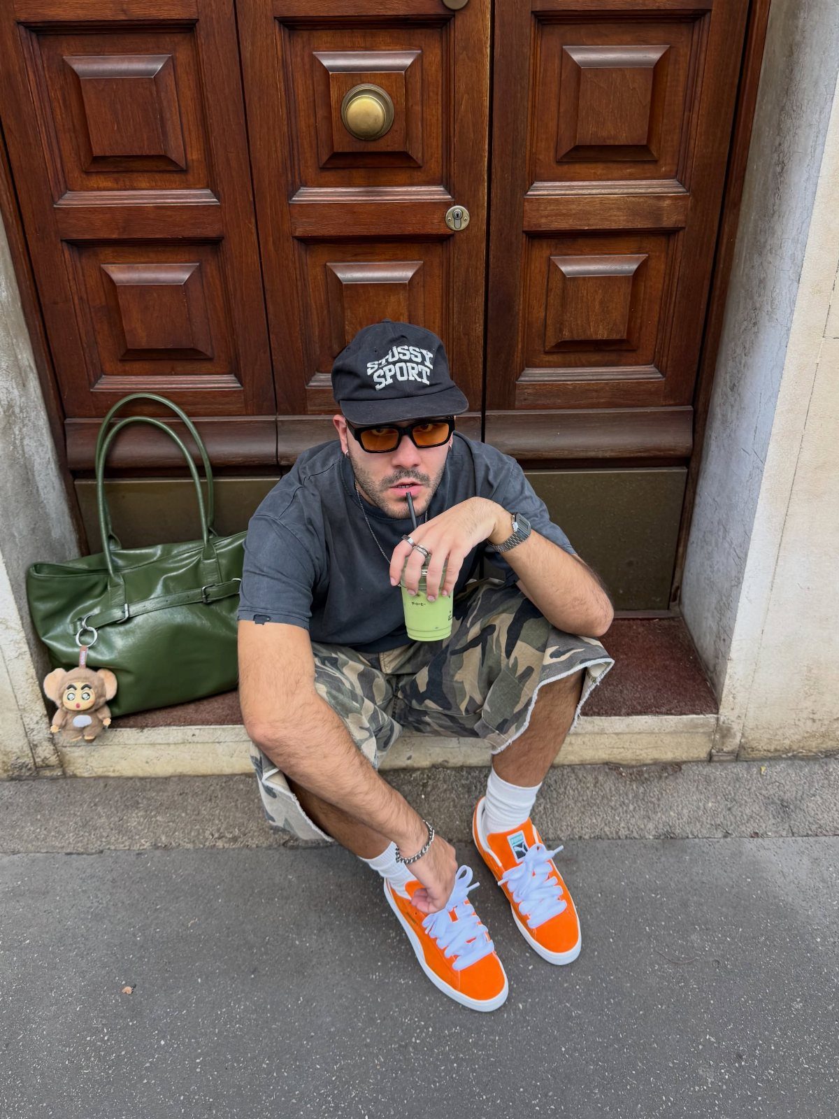 A man in camouflage shorts and a black cap wearing bright orange sneakers while sitting on a doorstep.