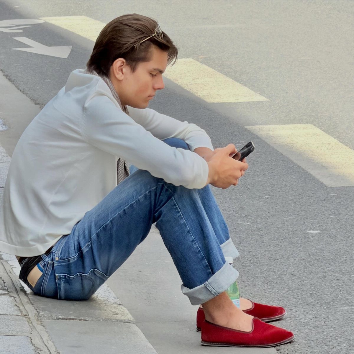 A young man in white shirt, blue jeans, and red loafers sits on a curb using a phone.