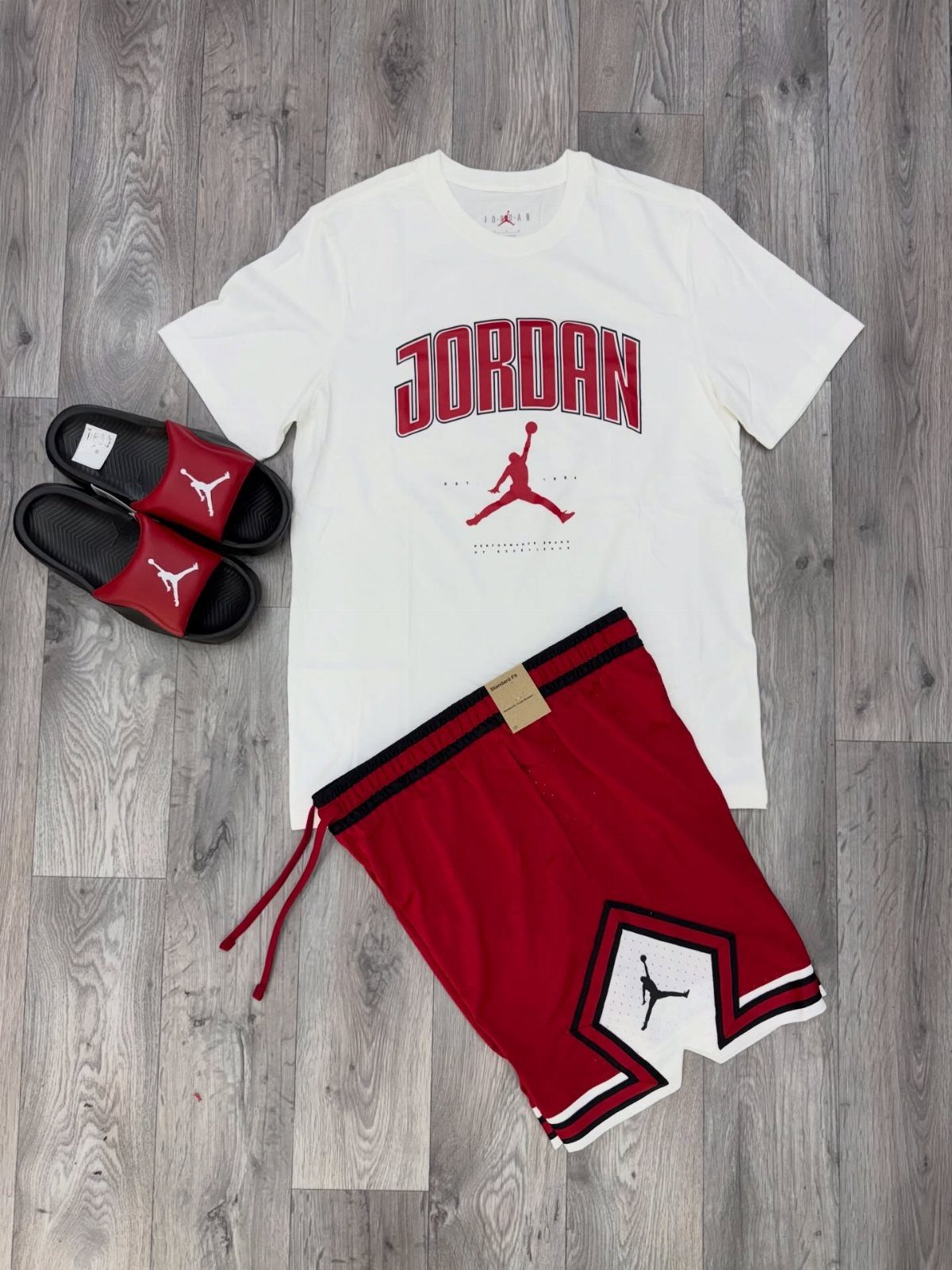 White Jordan t-shirt, red shorts, and red slides laid on a grey wooden floor.