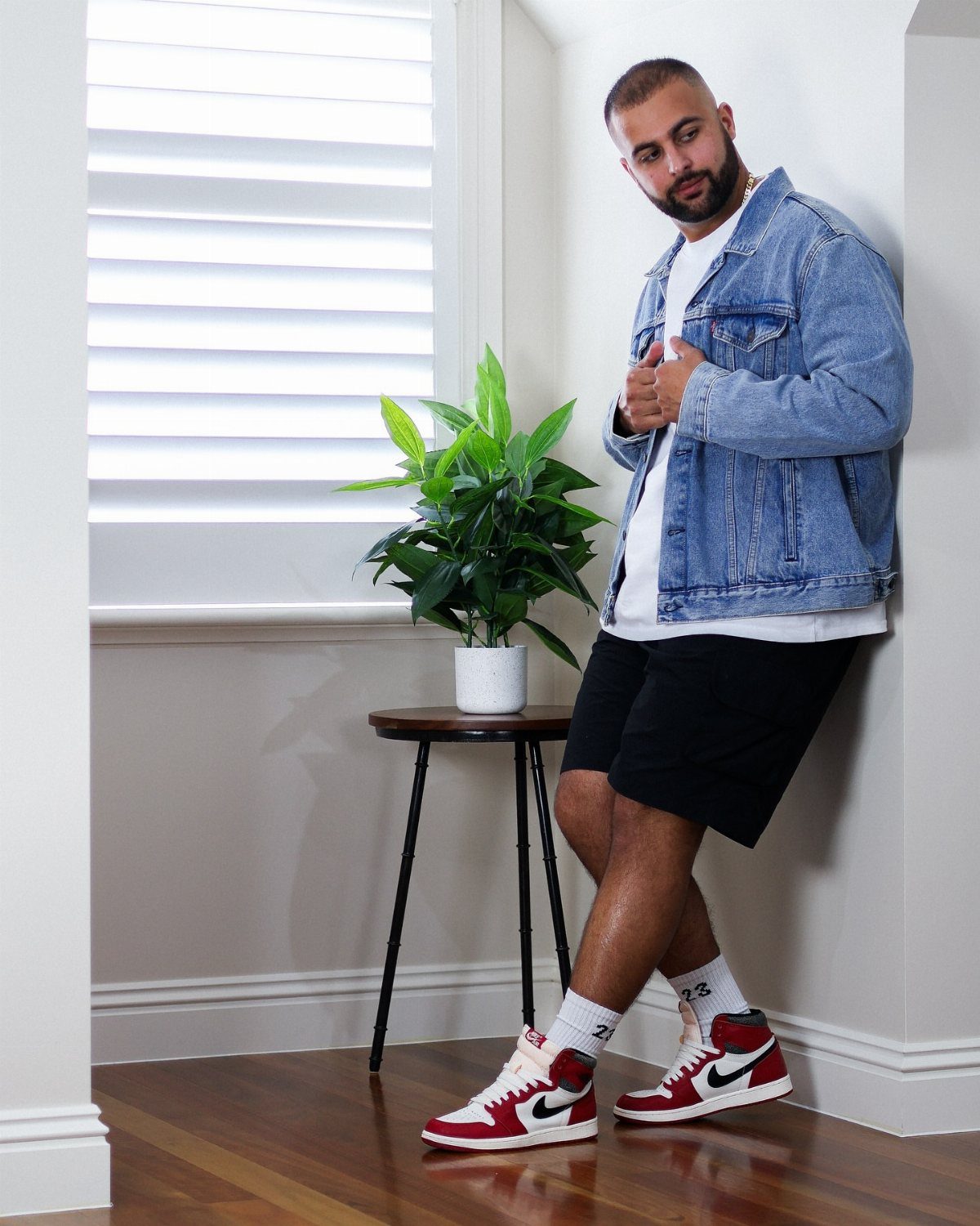 Man in a denim jacket, white t-shirt, black shorts, and red high-top sneakers leaning against a wall.
