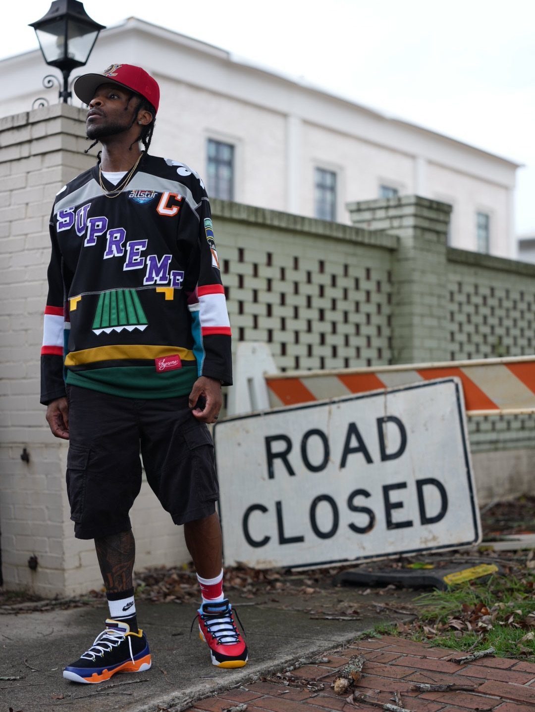 Man in a black Supreme jersey, black shorts, and colorful sneakers standing near a road closed sign.