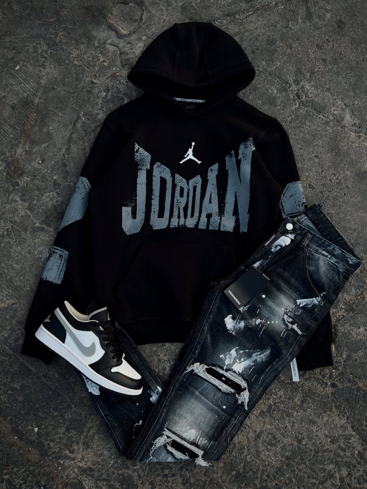 Black Jordan hoodie, distressed denim jeans, and black and white sneakers laid on concrete.