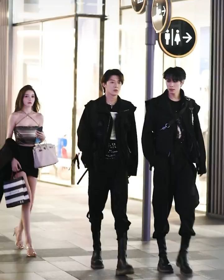Two men walking in black streetwear outfits on a city sidewalk near glass buildings.