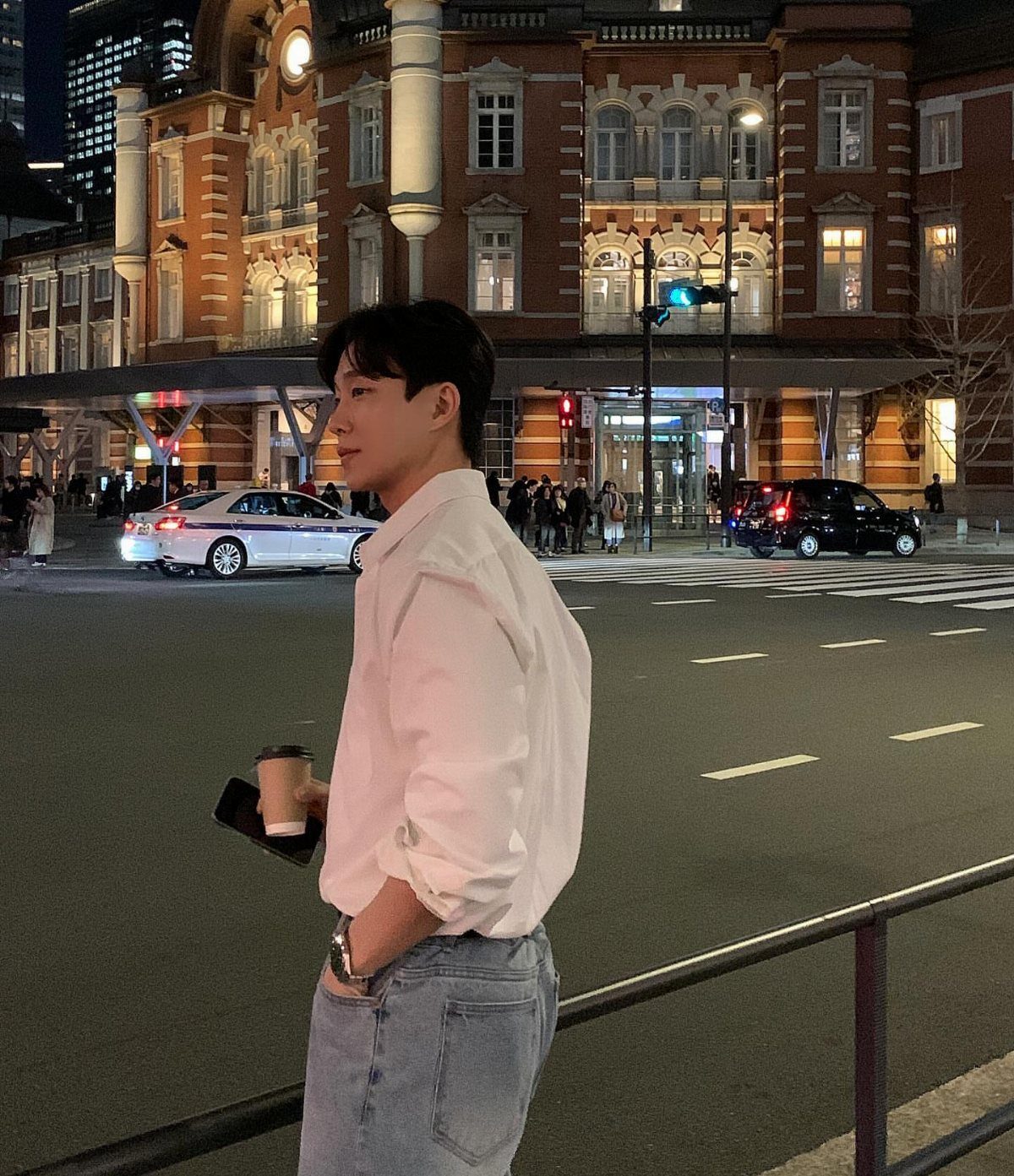A young man in a white shirt and light blue jeans standing on a city street at night.