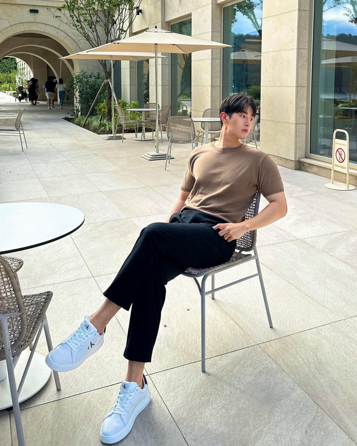 A man in a brown t-shirt and black trousers wearing bright white sneakers while sitting outdoors.