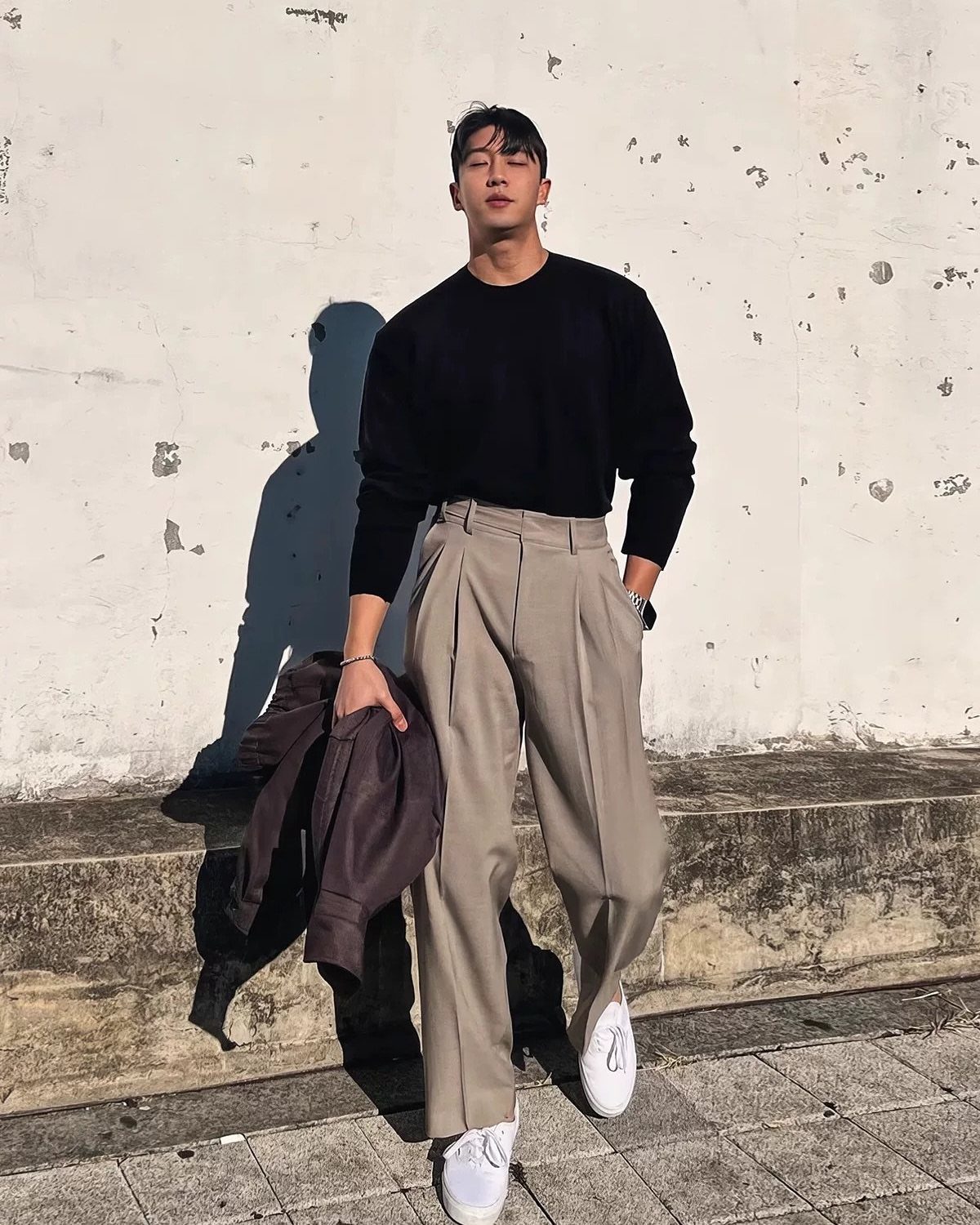 A man in a black sweater and beige pleated trousers standing against a white wall.