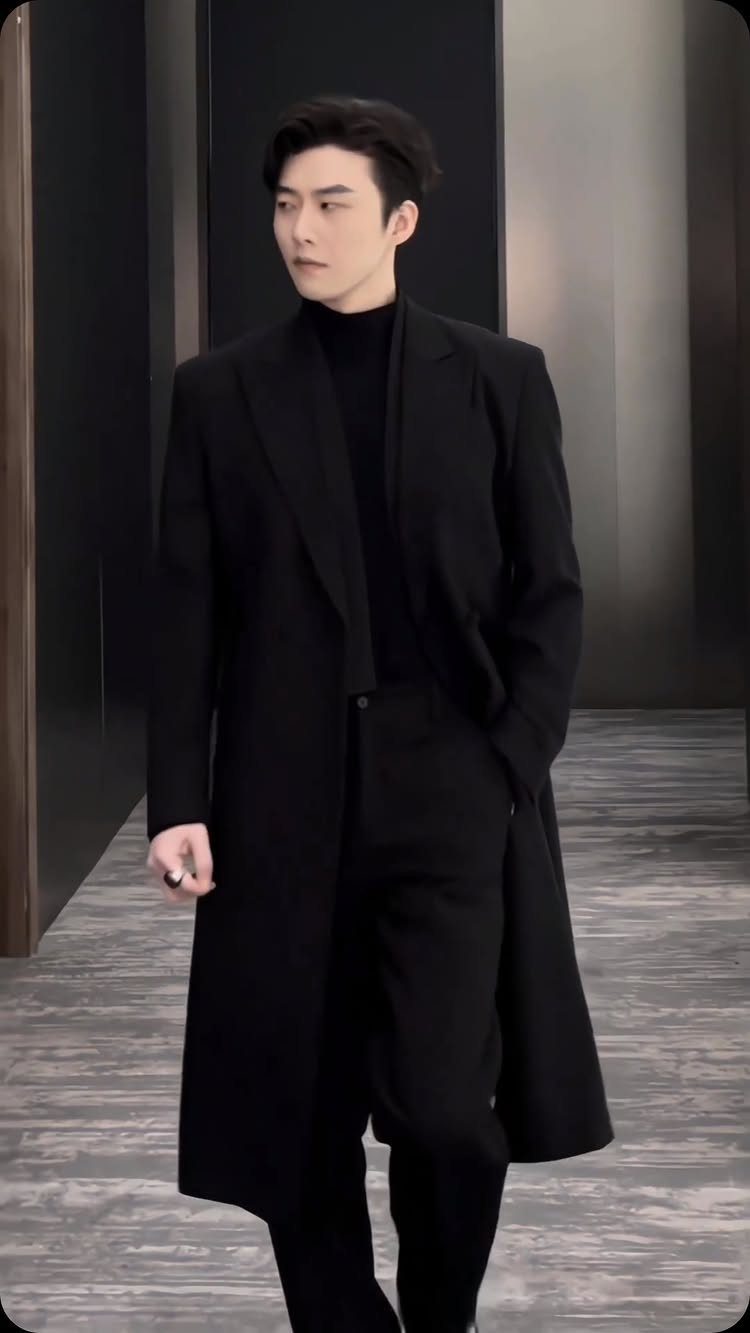 A young man in a black turtleneck and long black coat walking through a modern hallway.