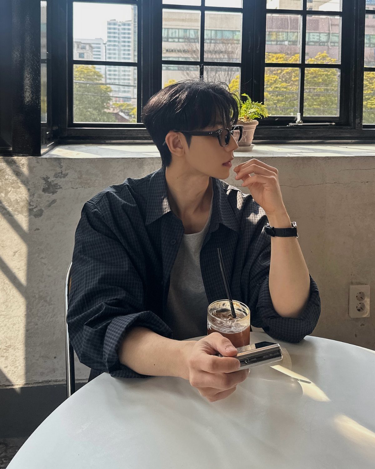 A young man wearing a dark patterned shirt and glasses sits at a cafe table.
