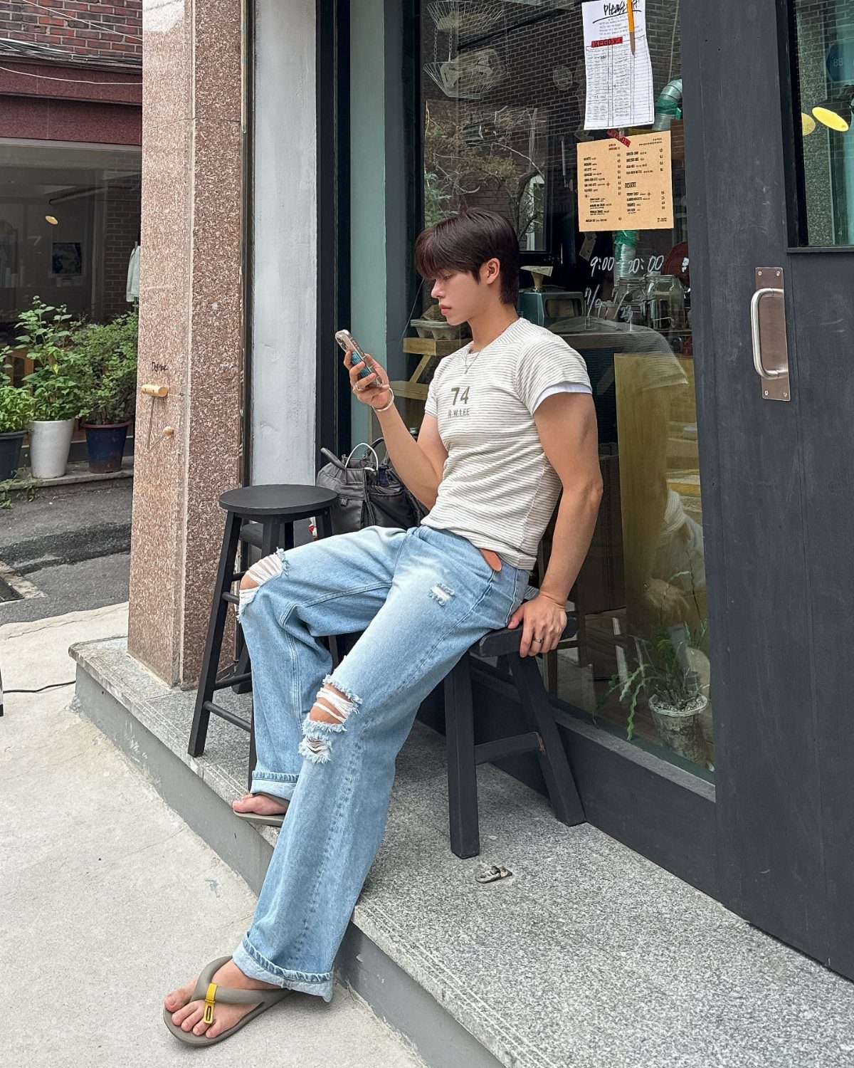 A young man in a white t-shirt and ripped light blue jeans sits on a stool outdoors.