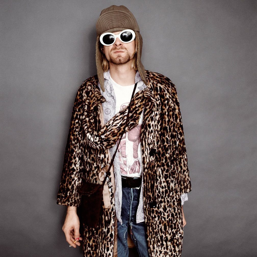 Man in leopard print coat, white sunglasses, and brown beanie posing against a grey background.