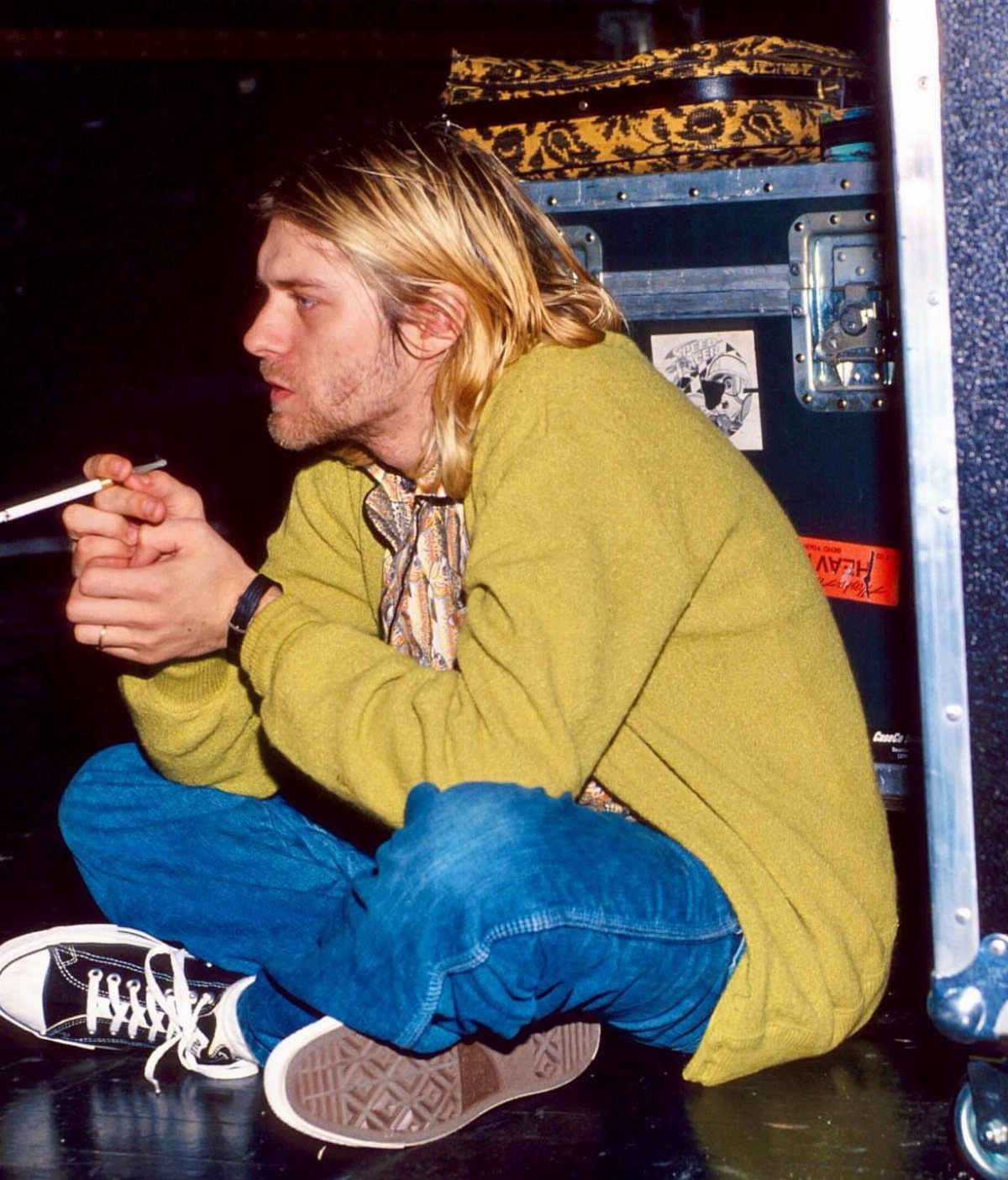 Kurt Cobain sitting on the floor wearing an oversized mustard yellow cardigan and blue jeans.