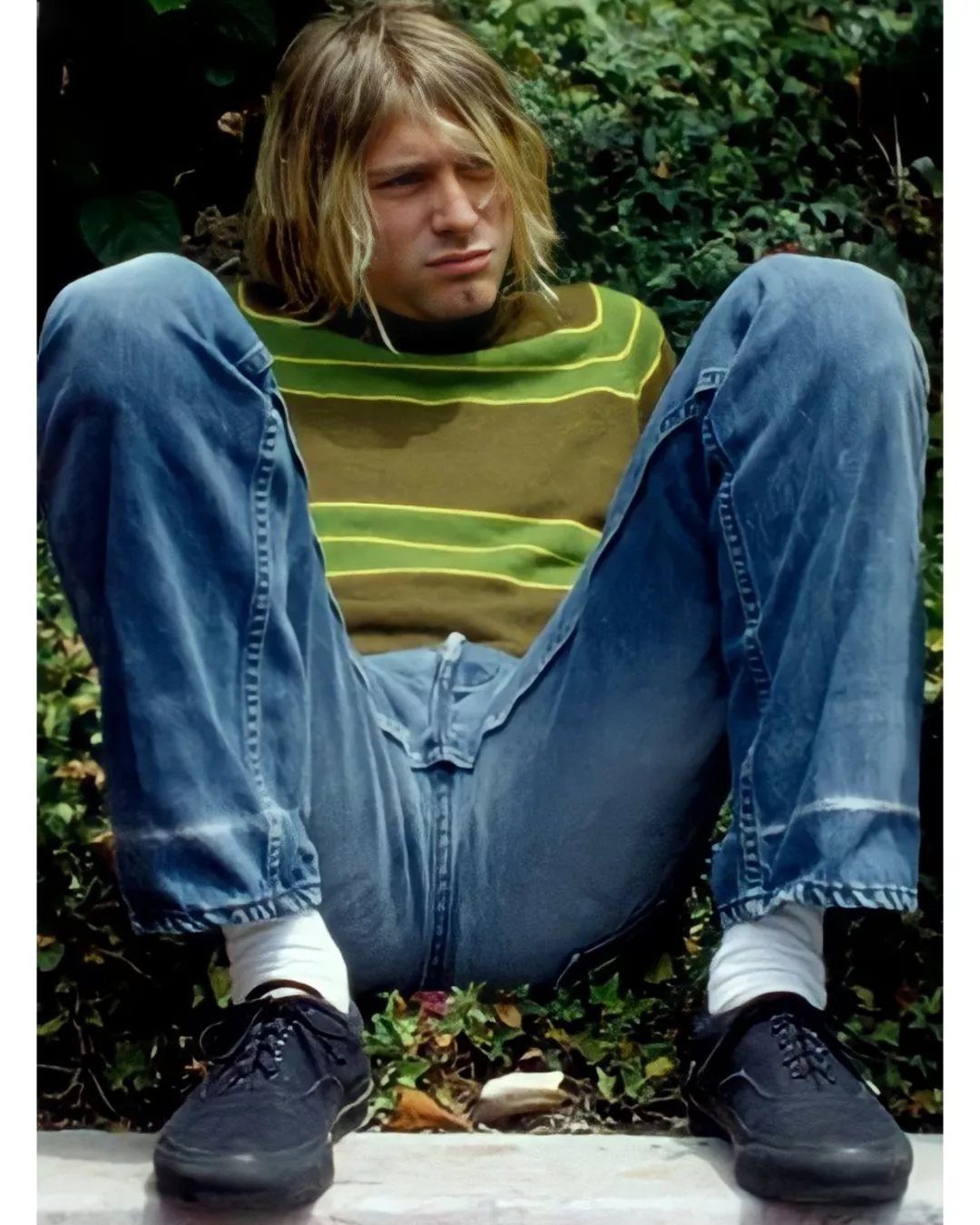 Man with long blonde hair sitting in blue jeans and black sneakers against green foliage.