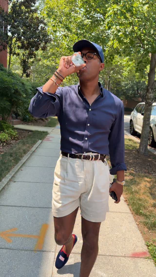 Man in navy shirt and cream shorts walking while drinking from a glass bottle.