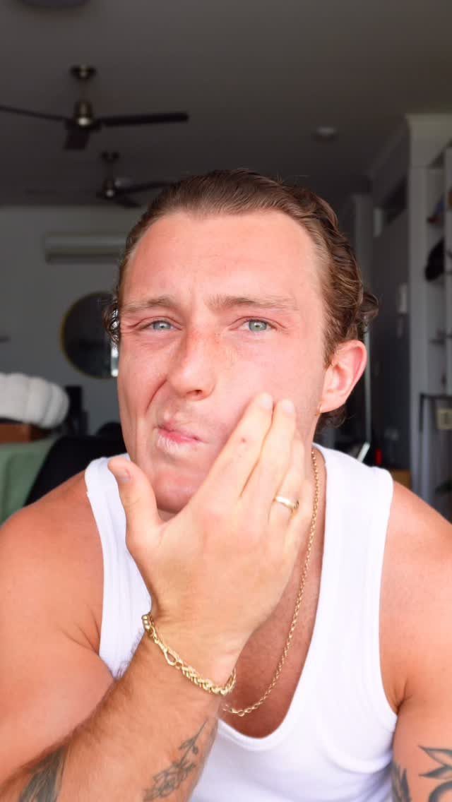 Man in a white ribbed tank top making a face while touching his cheek.