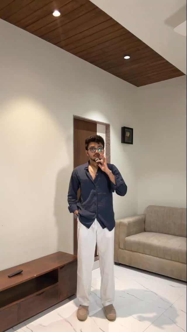 Man posing in a dark blue shirt and white linen pants in a modern living room.